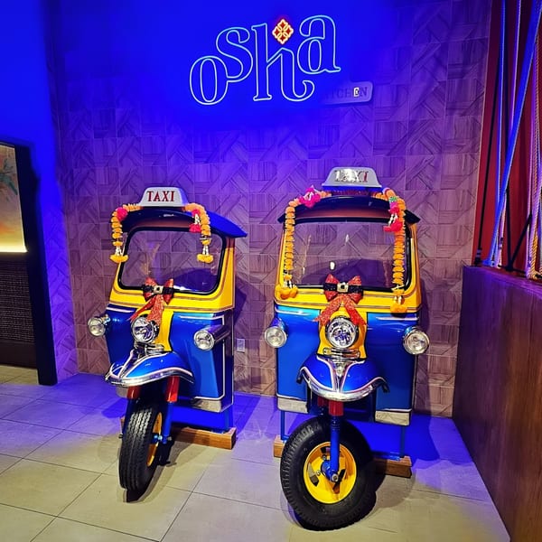 Two blue and yellow Osha Thai tuk-tuks with garlands and bows, under a neon sign.